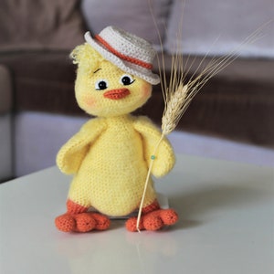 May include: A yellow crocheted chick wearing a white and orange hat holds a stalk of wheat. The chick has a friendly face with black eyes and a red beak.