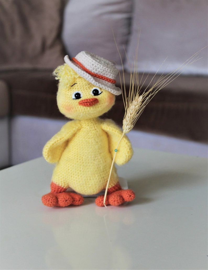 May include: A yellow crocheted chick wearing a white and orange hat holds a stalk of wheat. The chick has a friendly face with black eyes and a red beak.