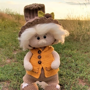 May include: A crocheted gnome doll wearing a brown hat with green leaves, a yellow vest, and brown pants. The gnome is standing on a dirt path with green grass.