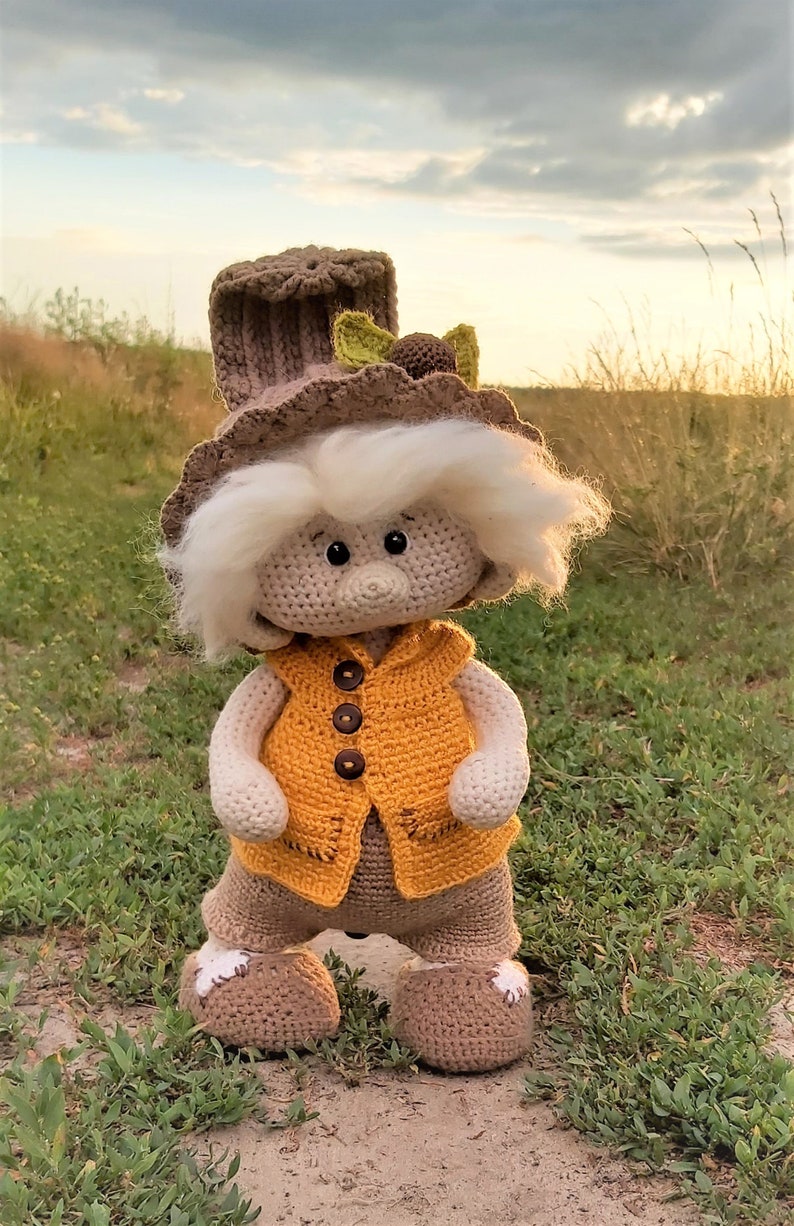 May include: A crocheted gnome doll wearing a brown hat with green leaves, a yellow vest, and brown pants. The gnome is standing on a dirt path with green grass.