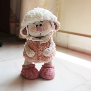 May include: A crocheted plush toy sheep with a white fluffy head, pink fur, and pink booties. The sheep is standing on a white tiled floor.