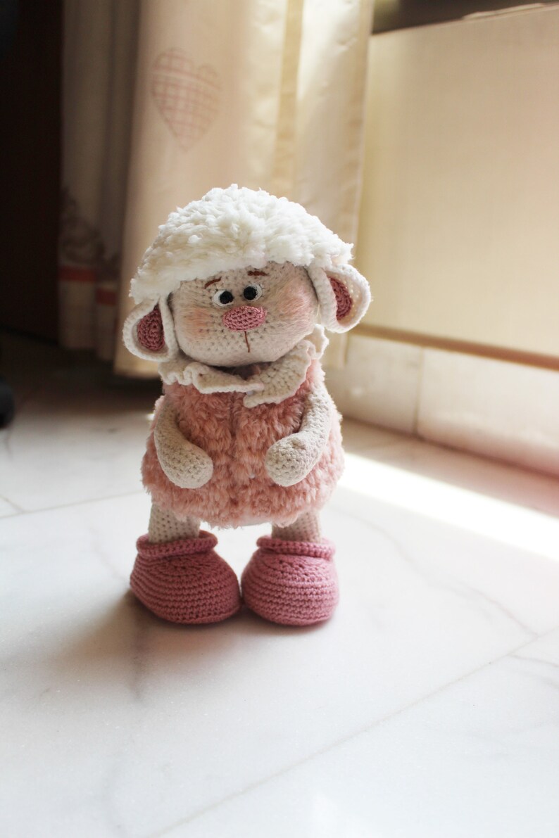 May include: A crocheted plush toy sheep with a white fluffy head, pink fur, and pink booties. The sheep is standing on a white tiled floor.