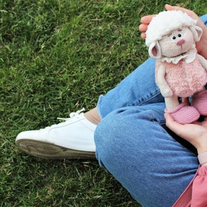 May include: A person wearing blue jeans and a pink shirt is holding a crocheted white and pink lamb toy. The lamb has a white face, pink body, and pink shoes.