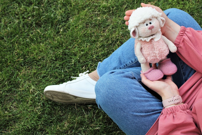 May include: A person wearing blue jeans and a pink shirt is holding a crocheted white and pink lamb toy. The lamb has a white face, pink body, and pink shoes.