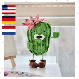 Peut inclure: Une figurine de cactus crochetée verte avec une fleur rose sur le dessus, des yeux noirs et blancs, un sourire brodé et des bottes crochetées marron. Le cactus a deux bras, dont un levé, et plusieurs épingles insérées sur le dessus de sa tête.
