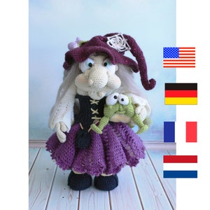 Op de afbeelding: Een gehaakte heksenpop met lang wit haar, die een paarse jurk, een zwart korset en een paarse hoed met een witte bloem draagt. Ze houdt een groene gehaakte kikker vast.