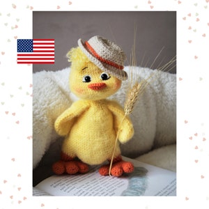 May include: A crocheted yellow chick wearing a beige hat with an orange band and holding a stalk of wheat. The chick is sitting on a book with a white fuzzy surface behind it.