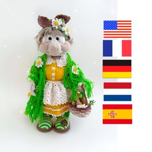 May include: A crocheted doll with a green shawl, yellow dress, and white apron. The doll is holding a basket of crocheted vegetables. The doll is wearing a brown headband with white flowers. The doll is standing on a white surface.