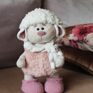 May include: A crocheted white lamb toy with pink fur, holding a smaller white crocheted lamb toy. The lamb is wearing pink crocheted booties.
