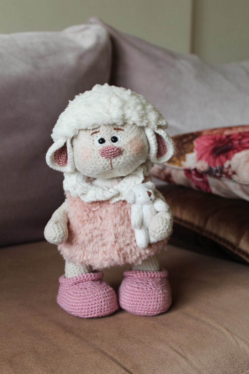 May include: A crocheted white lamb toy with pink fur, holding a smaller white crocheted lamb toy. The lamb is wearing pink crocheted booties.
