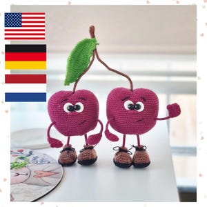 May include: Two crocheted cherry figures in deep magenta yarn with black bead eyes and stitched smiles. Each cherry has thin brown stems connected to a single green leaf, brown yarn shoes, and small arms, standing upright as a pair.