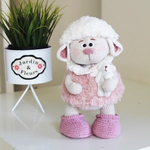 May include: A white crocheted lamb doll wearing a pink fuzzy dress and pink booties. The doll is holding a small white crocheted lamb in its arms. The doll is sitting on a white table.