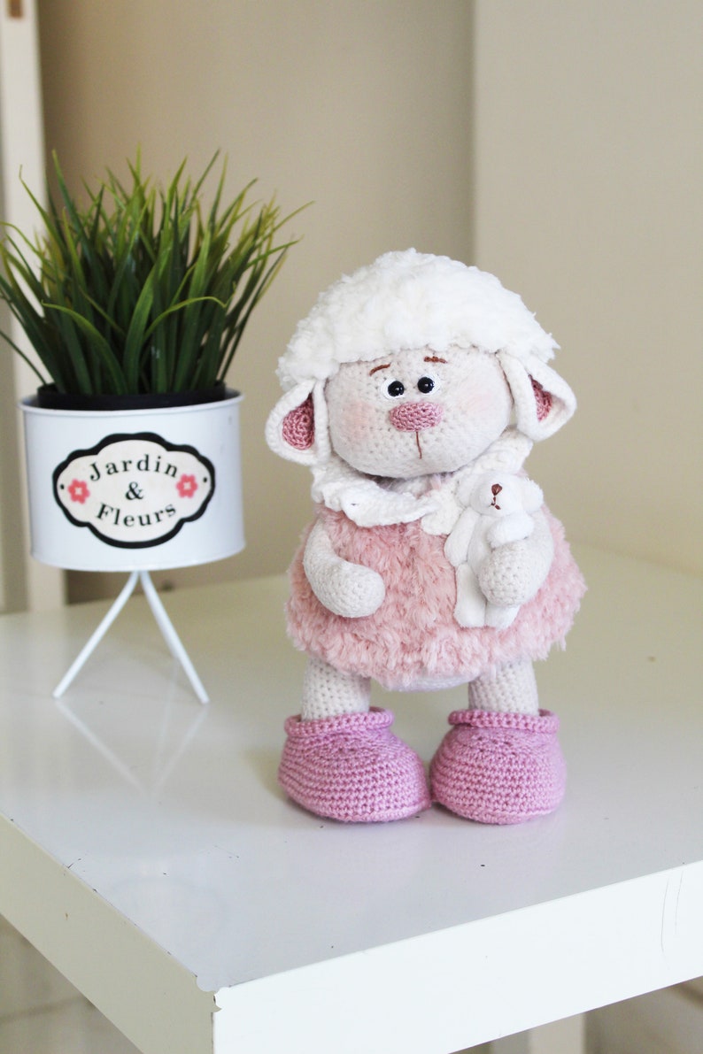 May include: A white crocheted lamb doll wearing a pink fuzzy dress and pink booties. The doll is holding a small white crocheted lamb in its arms. The doll is sitting on a white table.
