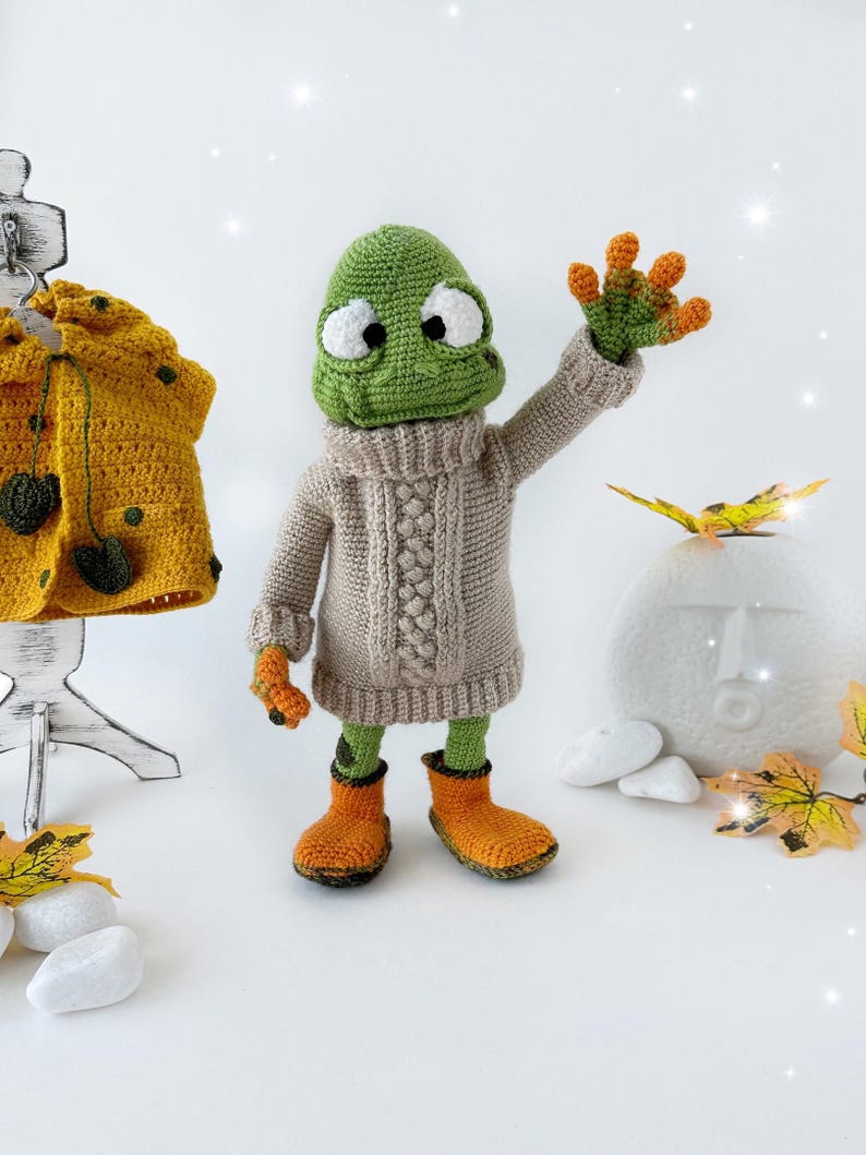 May include: A green crocheted frog wearing a beige knitted sweater and orange boots. The frog is standing on a white surface with fall leaves and white stones.