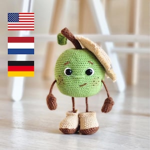 Op de afbeelding: Een handgemaakt, groen gehaakt appelkarakter met zwarte ogen, een lachende mond en bruine armen en benen. Het draagt beige laarzen en een bruine hoed. De appel heeft een bruine steel en een klein groen blad.