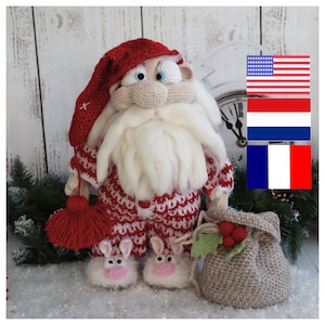 Puede incluir: Un muñeco de Papá Noel de crochet que lleva un pijama rojo y blanco y zapatillas de conejo. El muñeco sostiene una pequeña bolsa marrón con una rama de acebo verde y bayas rojas.