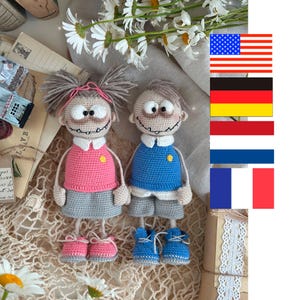 Könnte beinhalten: Zwei handgefertigte Häkelpuppen, ein Mädchen und ein Junge, stehen nebeneinander. Die Puppe des Mädchens trägt ein rosa Oberteil, einen grauen Rock und rosa Schuhe. Die Puppe des Jungen trägt ein blaues Oberteil, graue Shorts und blaue Schuhe. Beide Puppen haben lächelnde Gesichter.