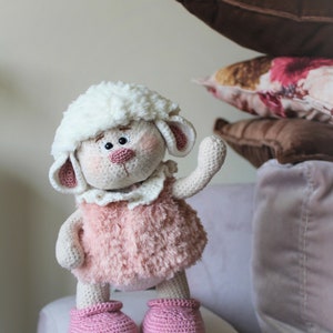 May include: A crocheted plush toy lamb with a white fluffy head, pink fur, and pink crocheted shoes. The lamb is standing on a light gray couch.
