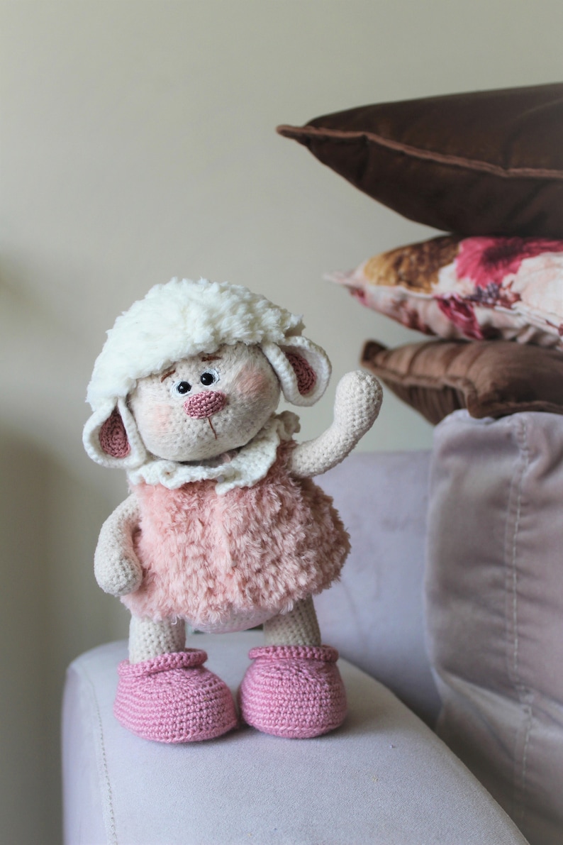 May include: A crocheted plush toy lamb with a white fluffy head, pink fur, and pink crocheted shoes. The lamb is standing on a light gray couch.