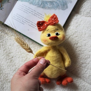 May include: A yellow crocheted chick toy with orange feet and a red and white crocheted bow. The chick is holding a wheat stalk in its beak.