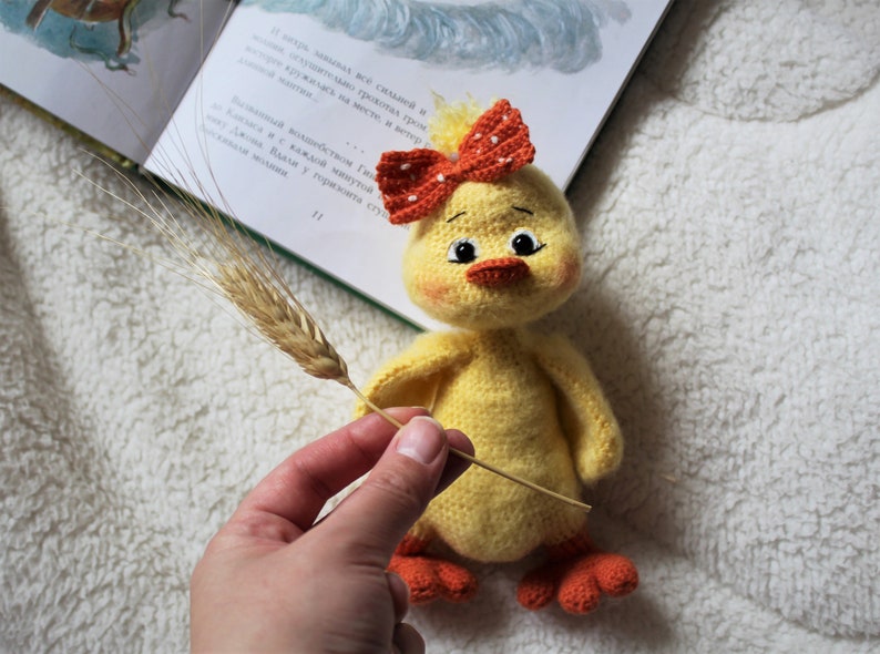 May include: A yellow crocheted chick toy with orange feet and a red and white crocheted bow. The chick is holding a wheat stalk in its beak.