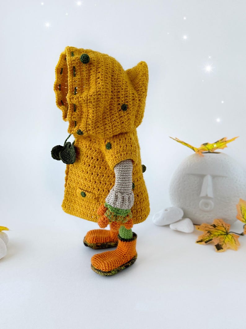 May include: A crocheted doll wearing a yellow raincoat with green accents, orange boots, and a gray knitted sleeve. The doll is standing on a white surface.