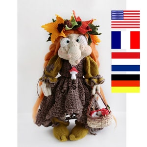 Puede incluir: Una muñeca de crochet con el pelo largo y rojo que lleva un vestido marrón con una chal verde y una corona de hojas de otoño. La muñeca lleva una cesta de setas rojas.