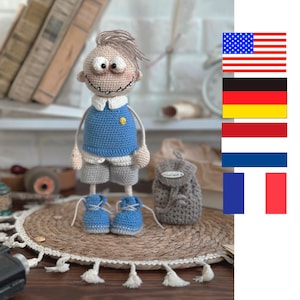 Könnte beinhalten: Handgefertigte Häkelpuppe eines Jungen mit blauem Pullover, grauen Shorts und blauen Schuhen. Die Puppe hat ein lächelndes Gesicht und hellbraunes Haar. Ein kleiner grauer Rucksack befindet sich neben der Puppe. Die Puppe ist etwa 25 cm groß.