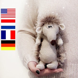 May include: A crocheted hedgehog plush toy with brown fur and large, black eyes. The toy is being held by a person's hand.