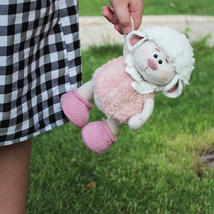 May include: A pink and white crocheted lamb toy with pink shoes. The lamb has a white fluffy head and a pink body. The toy is being held by a person.