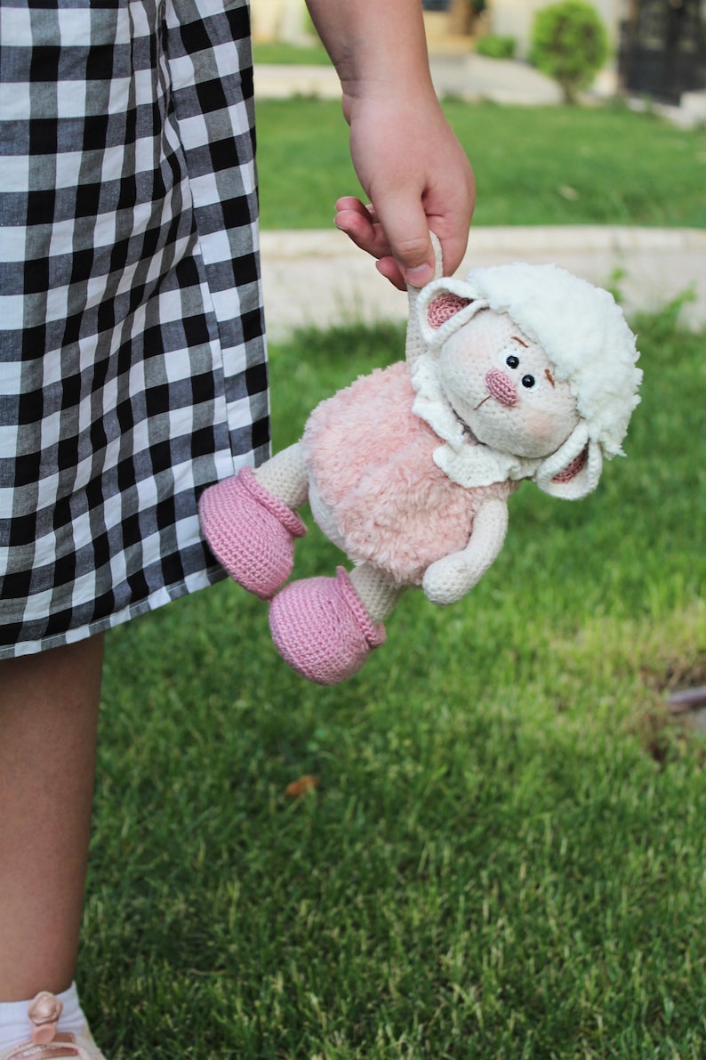 May include: A pink and white crocheted lamb toy with pink shoes. The lamb has a white fluffy head and a pink body. The toy is being held by a person.