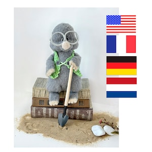 Może przedstawiać: Szary szydełkowy kret w zielonych okularach i zielonej kamizelce trzyma małą drewnianą łopatkę. Kret siedzi na stosie starych książek z kopą piasku przed sobą.