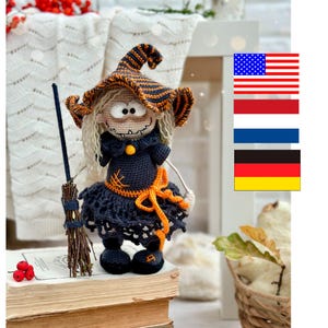 May include: A handmade crochet witch doll, featuring a black and orange striped hat and dress. The doll has a smiling face, blonde hair, and holds a broom. The witch is decorated with orange accents and a bow. The doll is sitting on a stack of books.