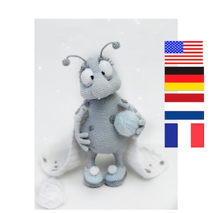 Puede incluir: Un juguete de amigurumi de color gris tejido a crochet con ojos grandes y una bola de lana en su mano. El juguete lleva botines tejidos a crochet de color blanco y tiene una manta de crochet blanca drapeada sobre su espalda.