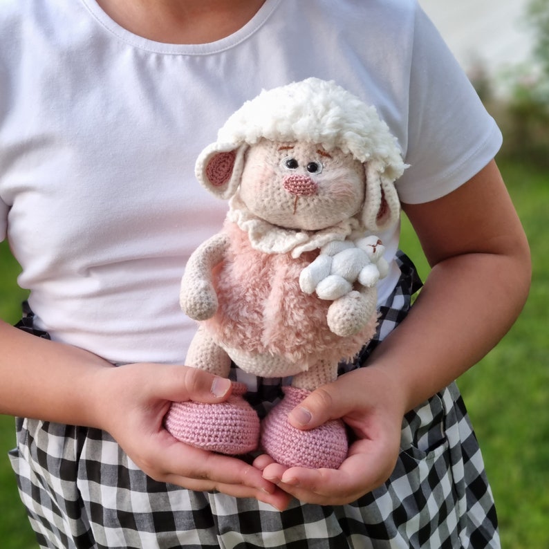 May include: A crocheted lamb toy with a pink and white fur coat, holding a smaller white lamb in its arms. The lamb is wearing a white crocheted collar and has a pink nose and brown eyes. The toy is being held by a person wearing a white shirt and a black and white checkered skirt.