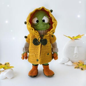 May include: A green crocheted frog wearing a yellow raincoat with a green hood and brown boots. The frog is standing on a white surface with fall leaves.