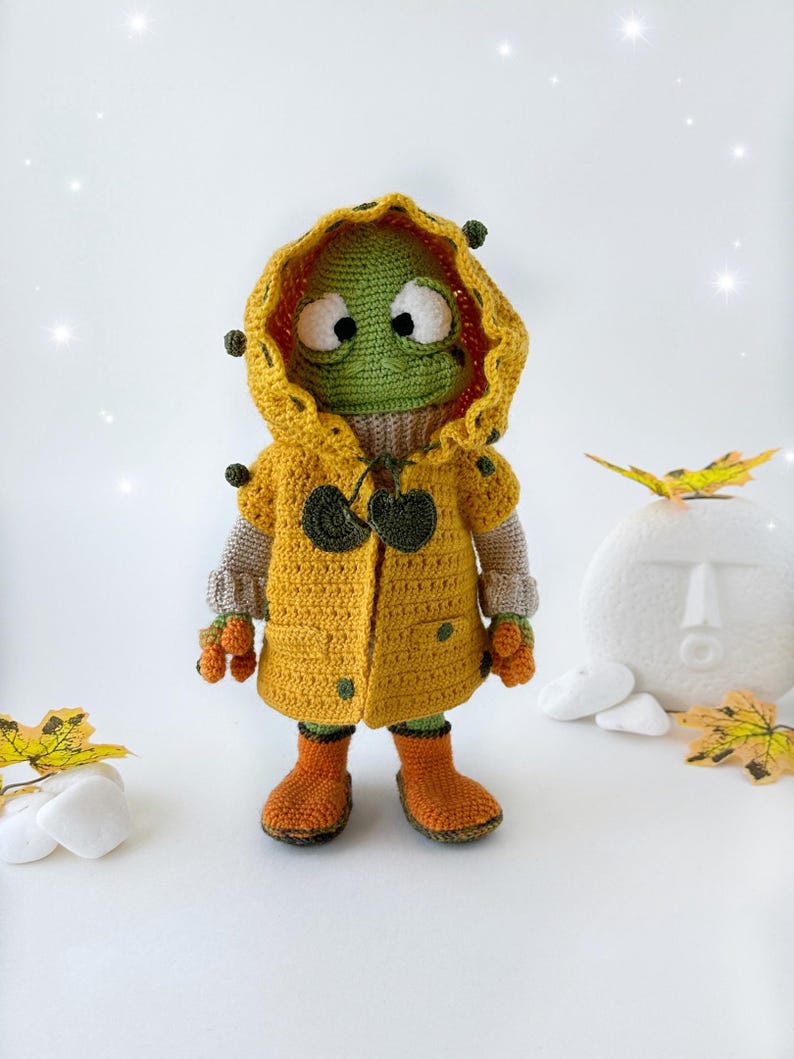 May include: A green crocheted frog wearing a yellow raincoat with a green hood and brown boots. The frog is standing on a white surface with fall leaves.