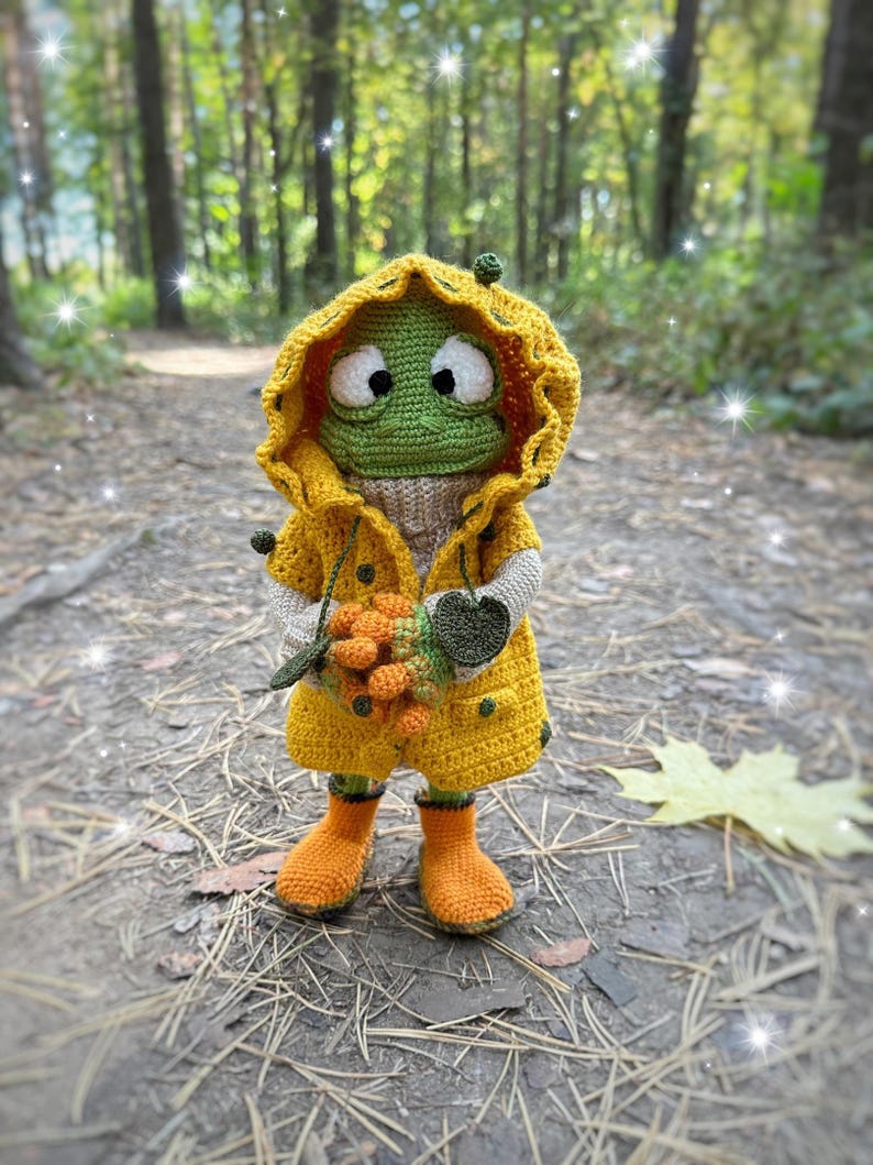 May include: A crocheted frog wearing a yellow raincoat and orange boots, holding a bunch of orange berries in a forest setting.