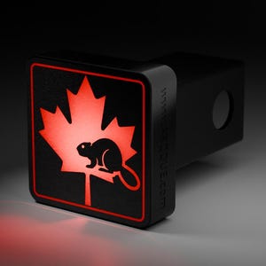 May include: Black trailer hitch cover featuring a red maple leaf and beaver design. The maple leaf and beaver are illuminated with red light. The hitch cover has a black border and a website address.
