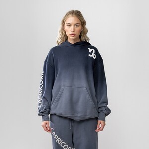 May include: A two-piece set of navy blue and gray ombre-dyed sweatsuit. The hoodie features the Capricorn symbol and the word "CAPRICORN" on the sleeve. The matching sweatpants also display "CAPRICORN". The outfit is worn with white sneakers.