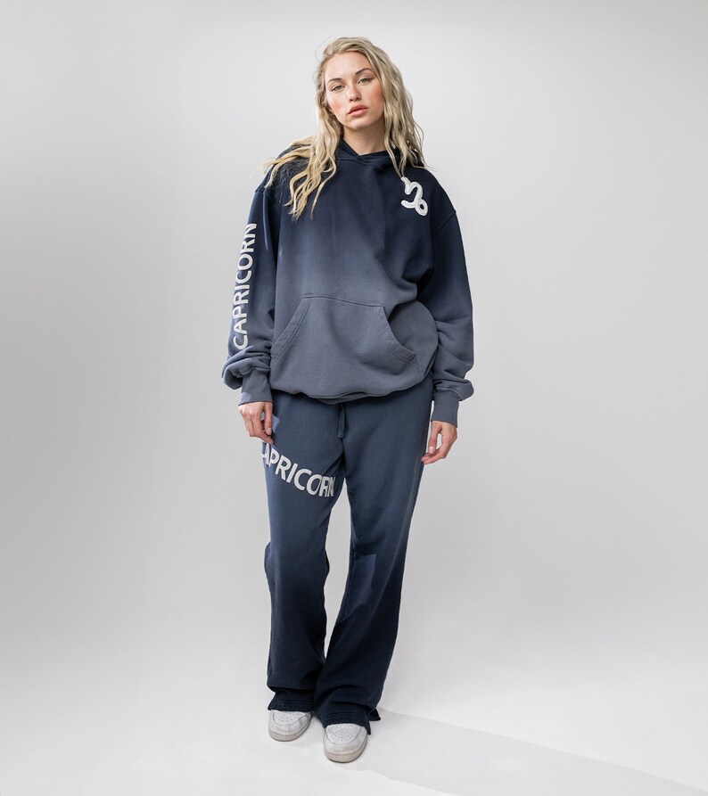 May include: A two-piece set of casual wear in a gradient blue hue. The set includes a hooded sweatshirt and matching sweatpants. The word "CAPRICORN" is printed in white on the left sleeve of the hoodie and on the left leg of the sweatpants. The Capricorn symbol is on the hoodie.