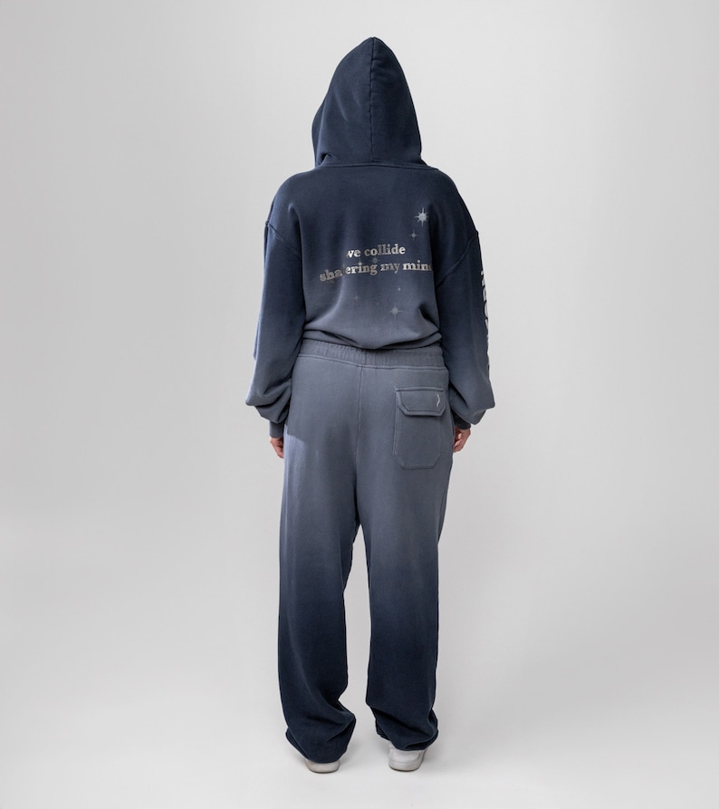 May include: A two-piece matching set of a hooded sweatshirt and sweatpants in a gradient blue color. The back of the hoodie has the text "we collide shattering my mind" with star graphics. The sweatpants have a single pocket on the back.