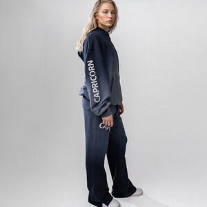 May include: A navy blue and gray ombre hoodie and matching sweatpants. The word "CAPRICORN" is printed vertically on the left sleeve of the hoodie. The word "CAP" is printed on the left pant leg. The outfit is worn with white sneakers.
