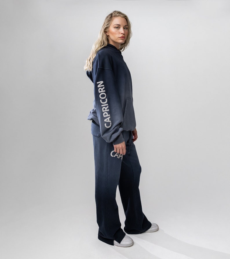 May include: A navy blue and gray ombre hoodie and matching sweatpants. The word "CAPRICORN" is printed vertically on the left sleeve of the hoodie. The word "CAP" is printed on the left pant leg. The outfit is worn with white sneakers.