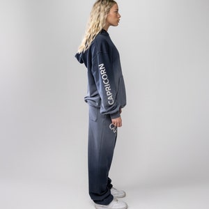 May include: A two-piece set of navy blue and gray-toned clothing. The hooded sweatshirt features the word "CAPRICORN" in white on the sleeve. The matching sweatpants also have the word "CAPRICORN" in white. The outfit is worn with white sneakers.