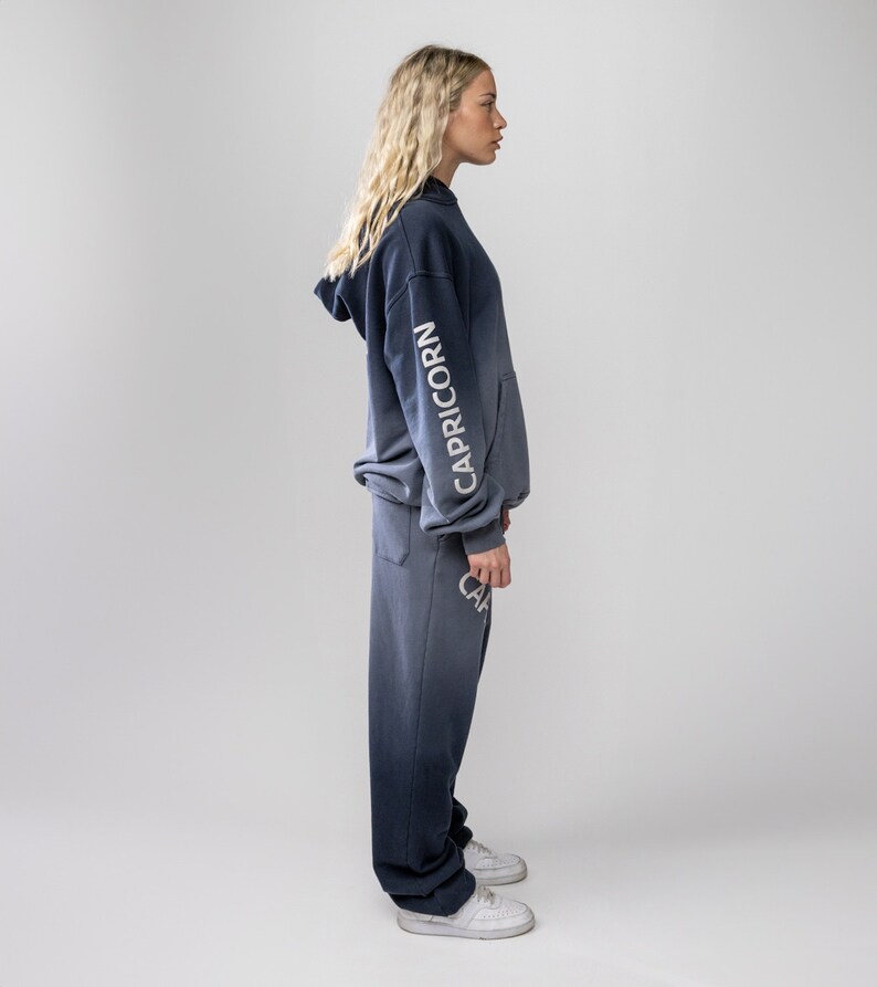May include: A two-piece set of navy blue and gray-toned clothing. The hooded sweatshirt features the word "CAPRICORN" in white on the sleeve. The matching sweatpants also have the word "CAPRICORN" in white. The outfit is worn with white sneakers.