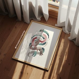 May include: A framed art print featuring a stylized illustration of a dragon with intertwined body. The dragon's body is a mix of white, pink, and teal colors. The frame is a light wood color, and the artwork is displayed on a wooden floor.