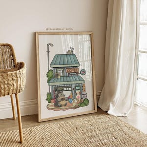 May include: Framed illustration of a coffee shop with a teal roof and the word "COFFEE" in a sign. A cartoon character is seated outside the shop. The artwork is in a light wooden frame and is leaning against a wall.