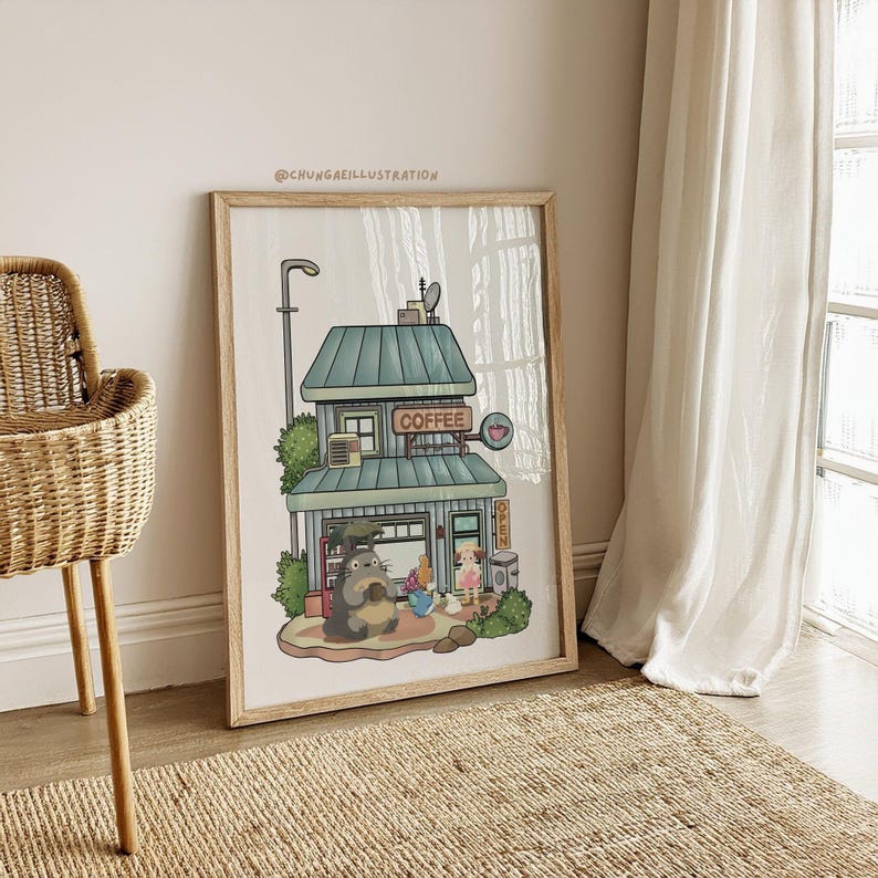 May include: Framed illustration of a coffee shop with a teal roof and the word "COFFEE" in a sign. A cartoon character is seated outside the shop. The artwork is in a light wooden frame and is leaning against a wall.