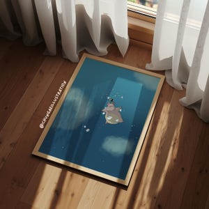 May include: A framed art print with a cartoon hippo floating in a blue space with clouds. A small bird sits on the hippo's head. The frame is light brown, and the artwork is displayed on a wooden floor. The artist's name is visible.